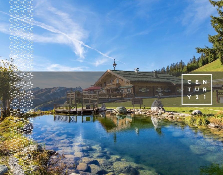 Rustic cabin with a wooden deck overlooking a clear pond in a scenic mountain landscape under a bright blue sky